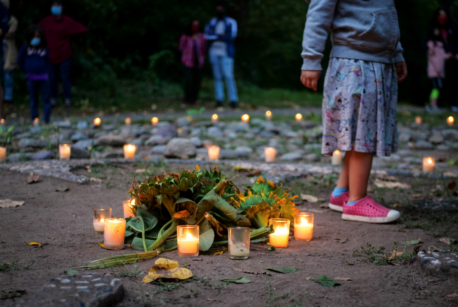A pile of sunflower heads on the ground, surrounded by lit tealights, with more lit tealights and people in the background